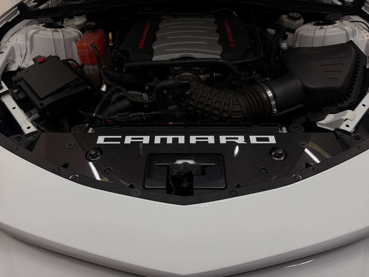 
                  
                    Close-up of the open front hood of a white Camaro, showing the engine bay with a black Camaro-branded engine cover and carbon-fiber-style panel.
                  
                