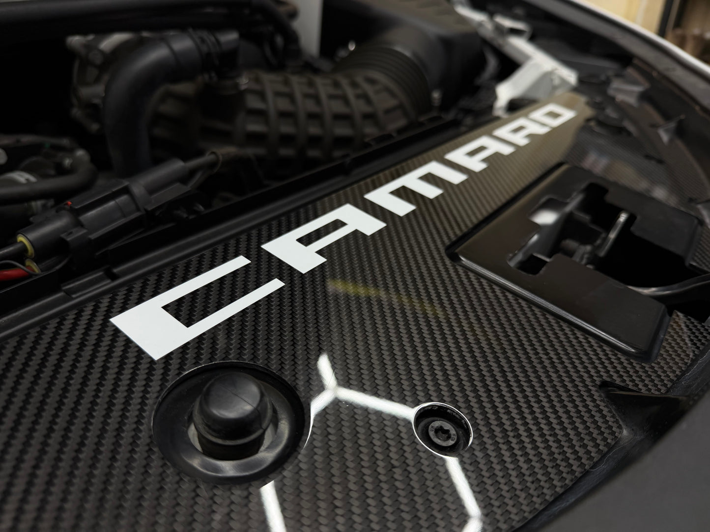 
                  
                    Close-up of a carbon-fiber-look engine cover with large white CAMARO lettering, photographed inside an open car hood.
                  
                