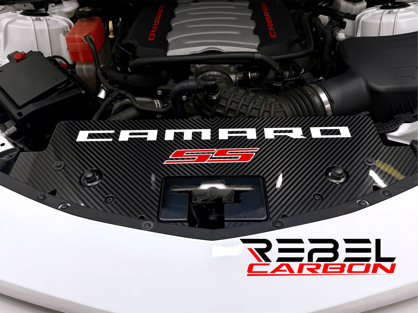 
                  
                    A carbon fiber engine cover lip installed on a Camaro SS vehicle, featuring a black carbon fiber pattern with white and red Camaro branding and a Rebel Carbon logo overlay.
                  
                