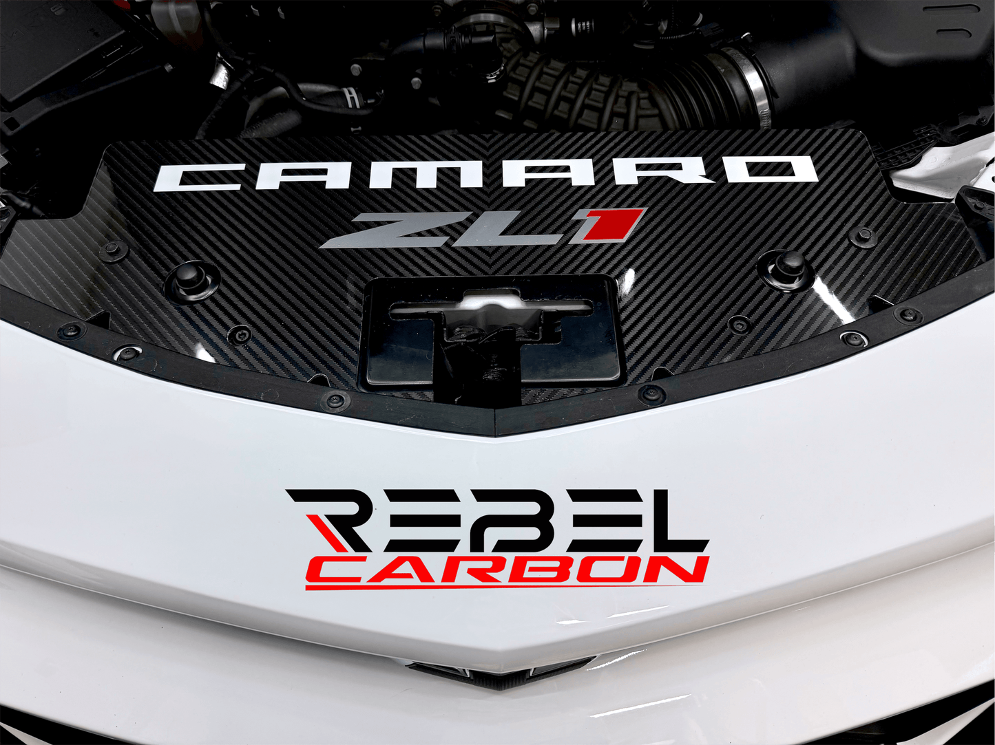 
                  
                    A close-up of a white car featuring a black carbon-fiber-style decal radiator cover with the words 'CAMARO ZL' in white and red, plus a lower decal reading 'REBEL CARBON' in black and red.
                  
                