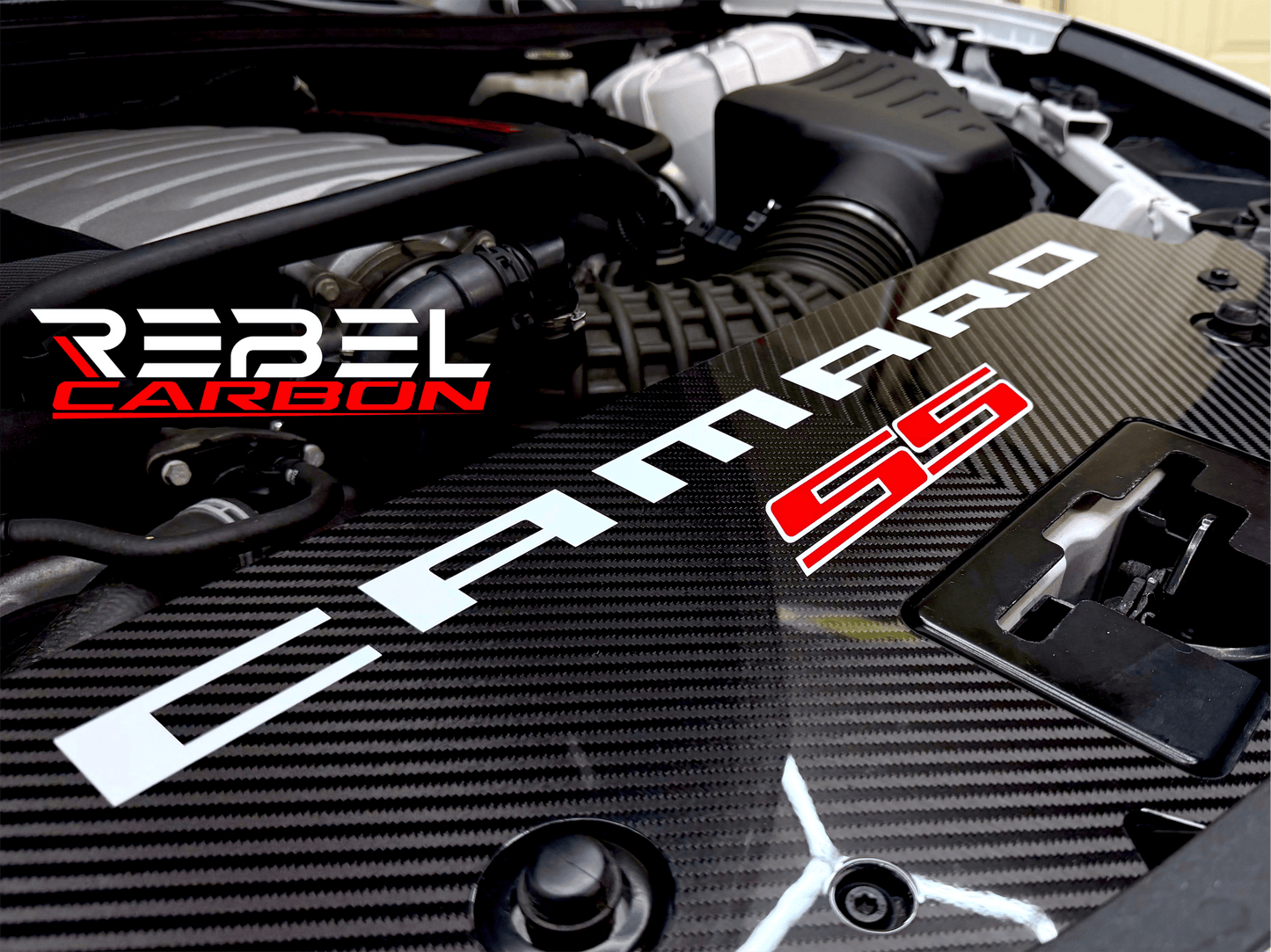
                  
                    A carbon fiber radiator cover on a Camaro SS with a Rebel Carbon logo overlay, shown in a close-up of the vehicle's engine bay.
                  
                