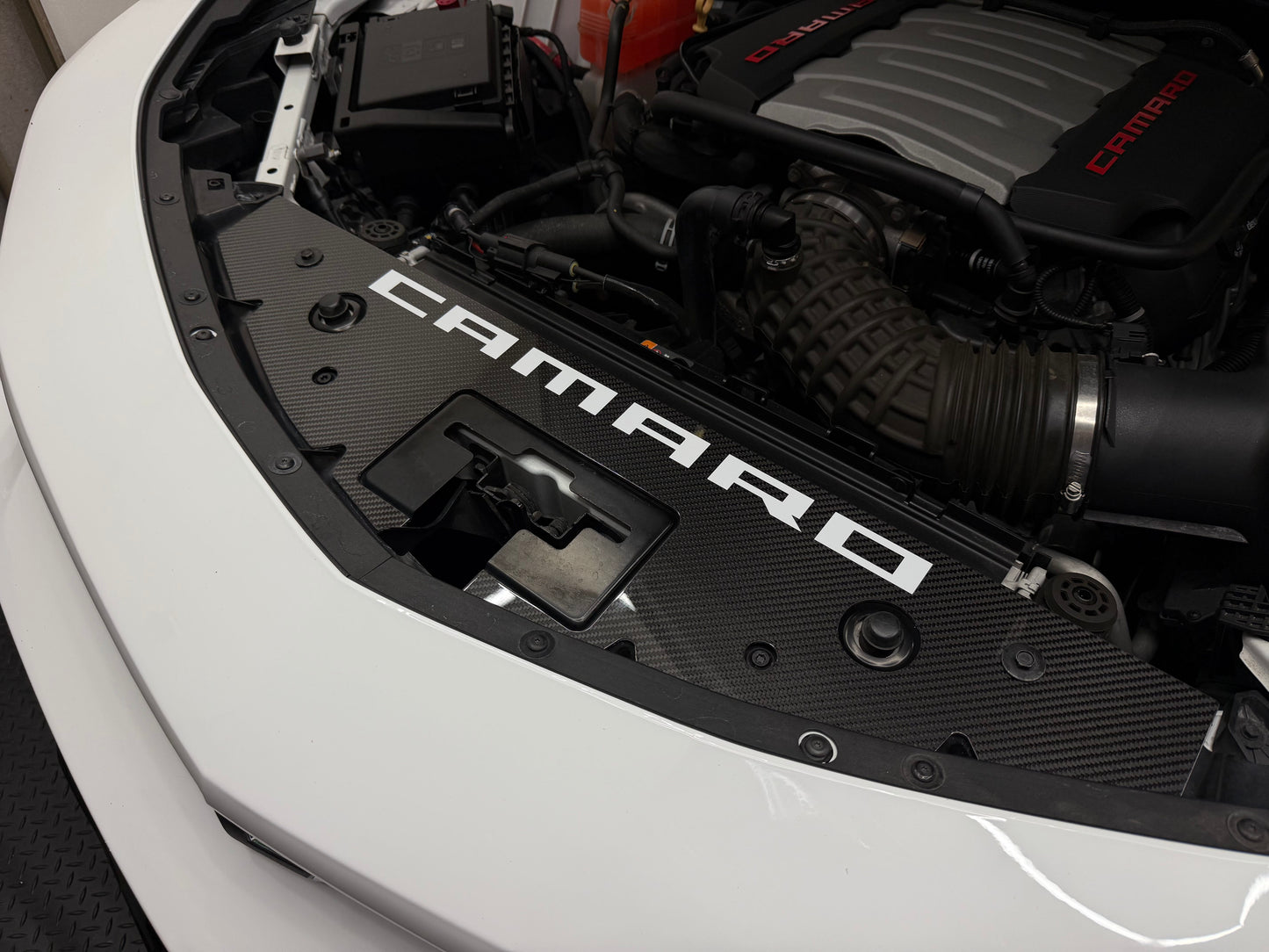 
                  
                    A close-up of a black carbon-fiber-style engine cover with large white CAMARO lettering, installed in the open hood of a white vehicle. The engine bay also shows a black Camaro engine cover with red lettering.
                  
                